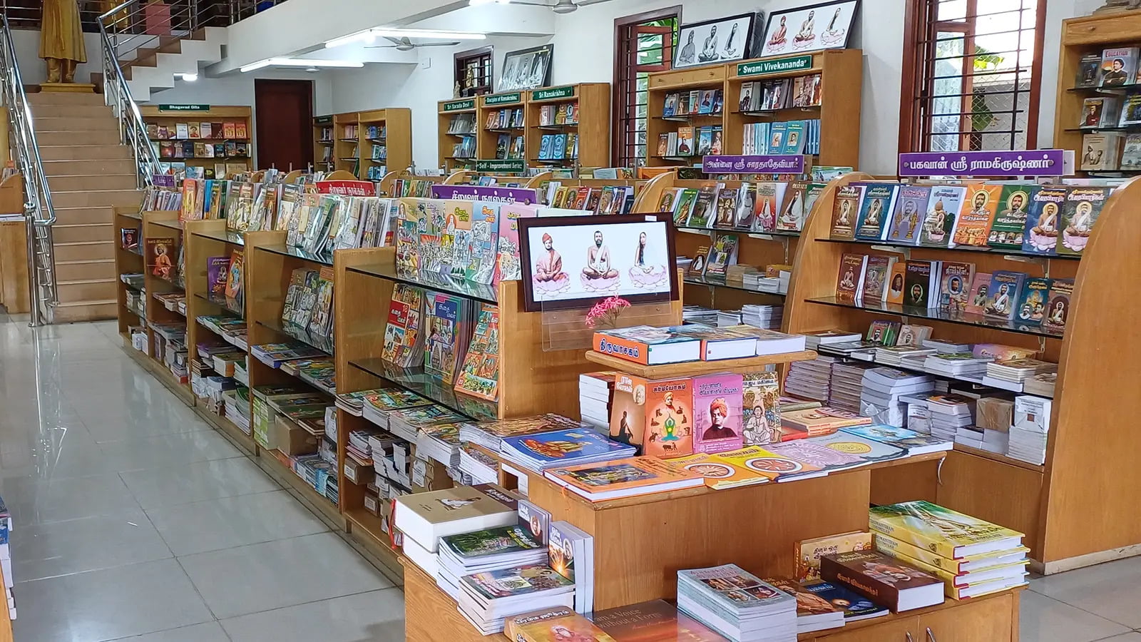 Interior view of a well-lit bookstore of Ramakrishna Math Madurai, with a large collection of books on white shelves and a staircase in the background